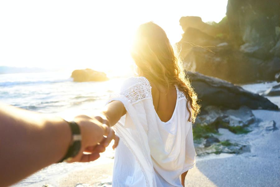 girl walking towards the ocean on a beach with the sun while a guy is holding her hand